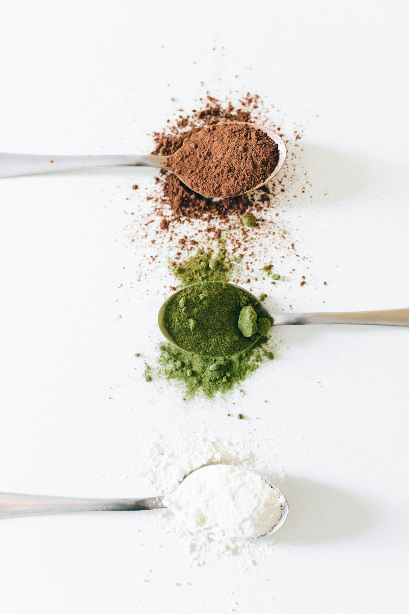 Flat lay of spoons with brown, green, and white powders on a white background.