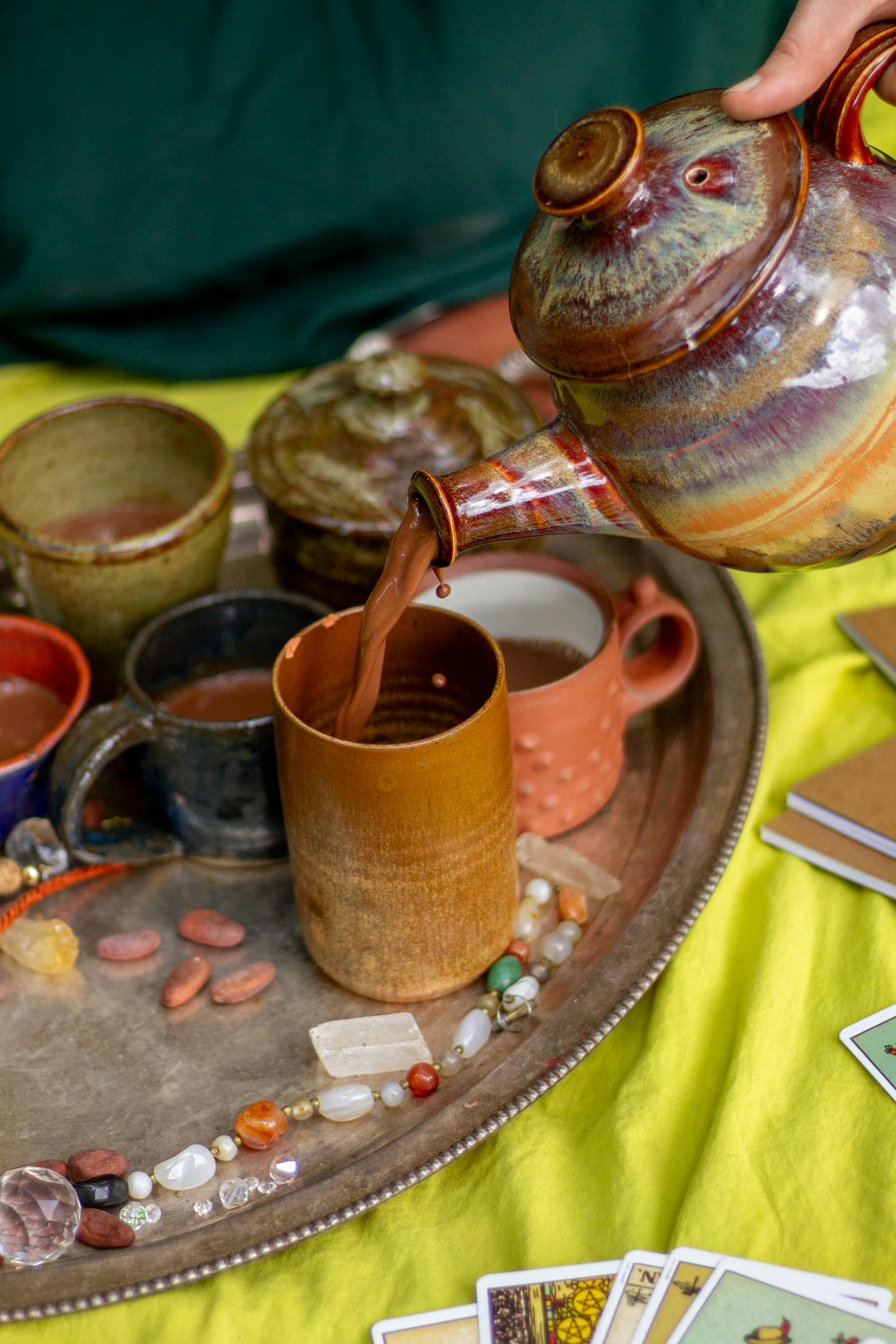 Hand pouring tea from pottery teapot surrounded by crystals and tarot cards.